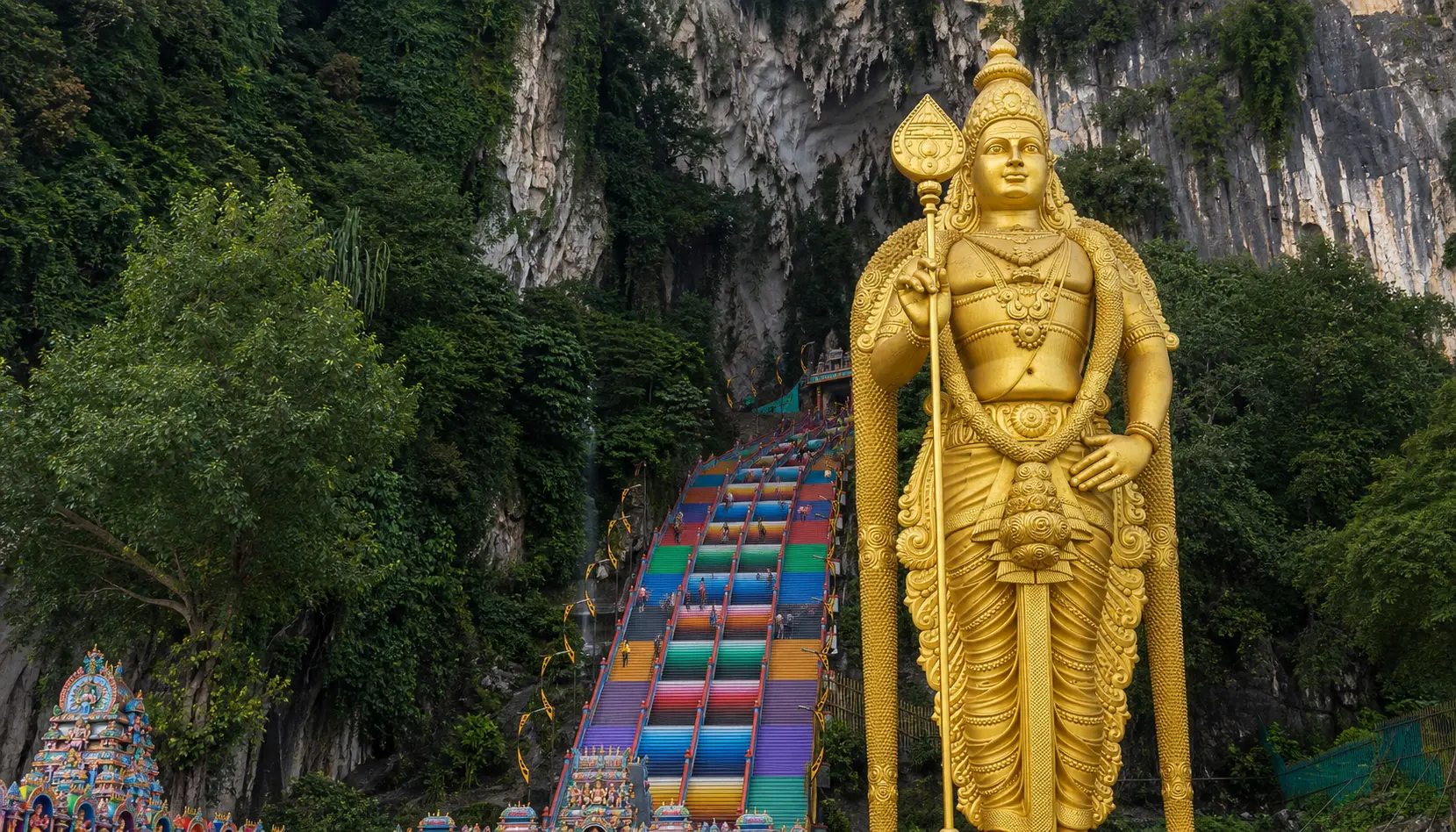 The rainbow-painted staircase at Batu Caves in Malaysia rising to a limestone cave entrance, with the tall gilded Murugan statue standing at the base